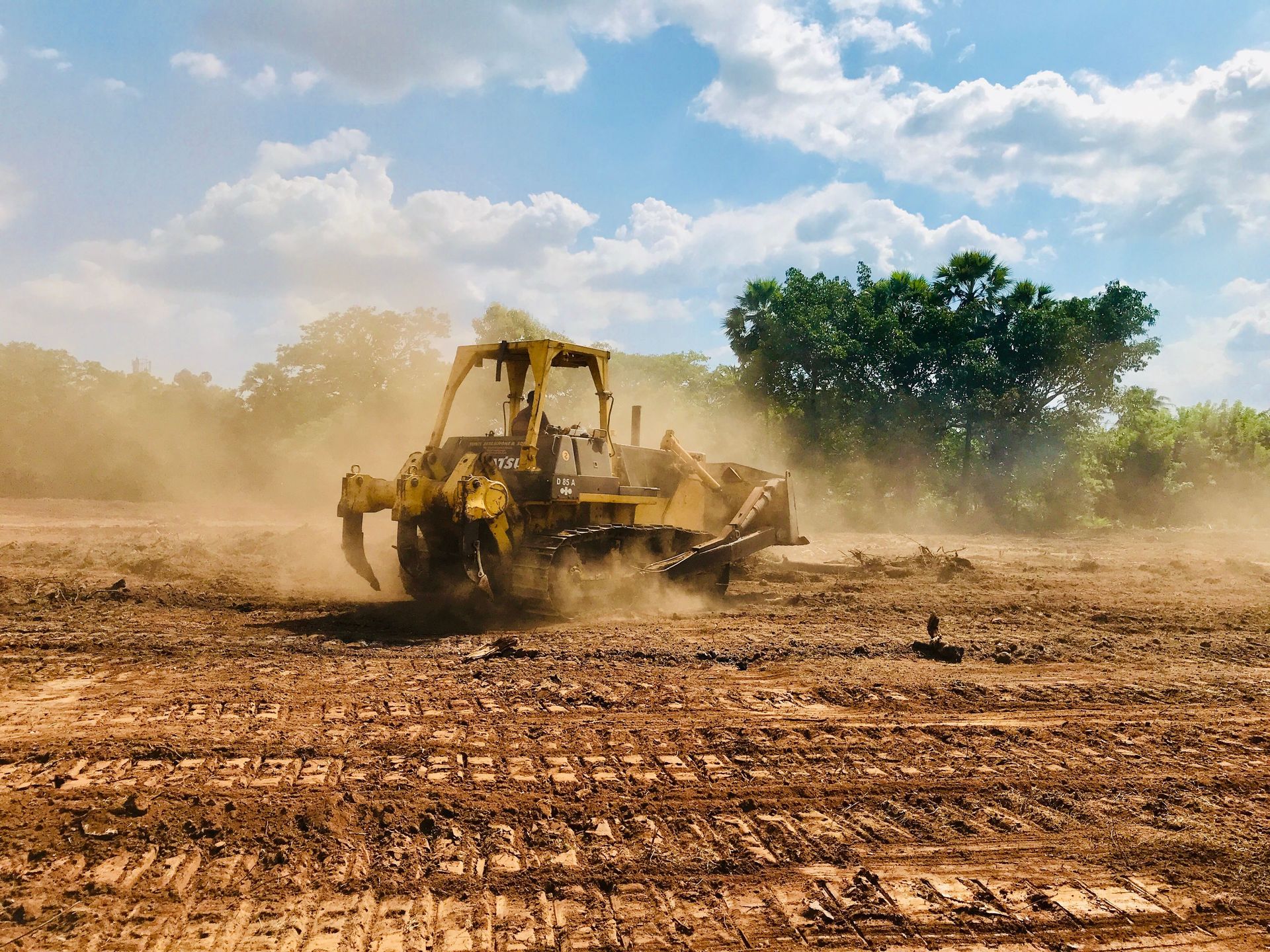 A powerful bulldozer works on a dusty, dirt field under a bright blue sky with cloud cover and trees in the background.